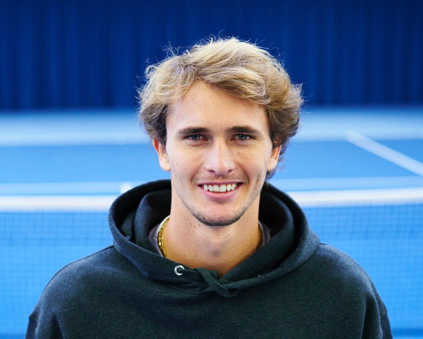 Porträt von Alexander Zverev im Kapuzenpullover vor einem blauen Tennisplatz